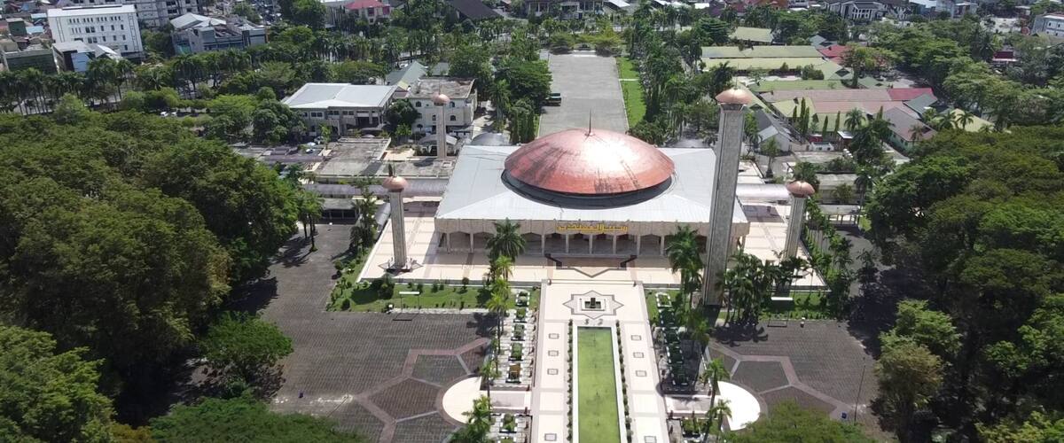 An aerial view of a large mosque with a prominent dome, surrounded by greenery and urban buildings.