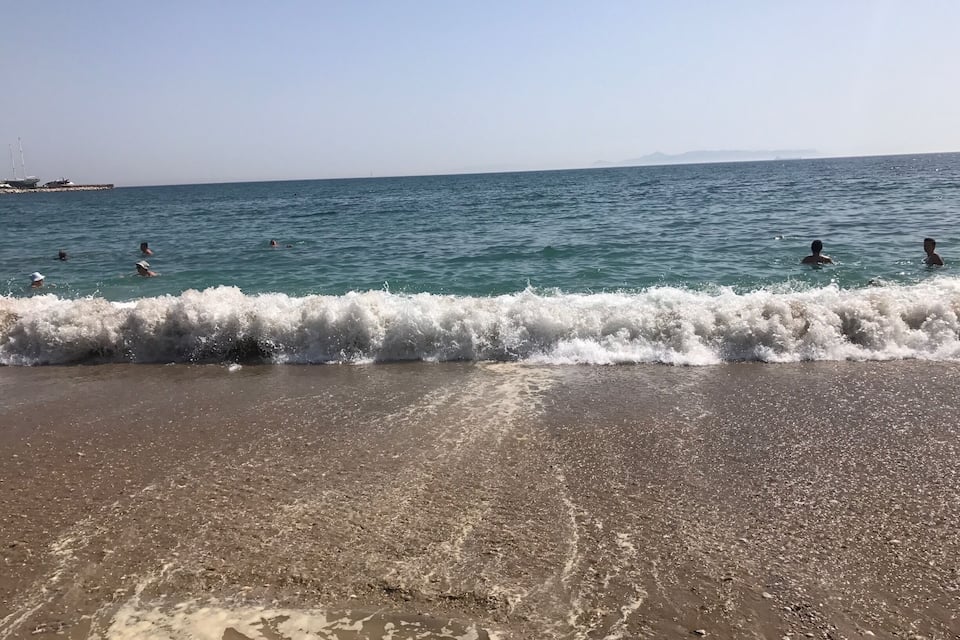 Uma praia com ondas quebrando na costa, pessoas nadando na água e uma linha de costa distante com montanhas.