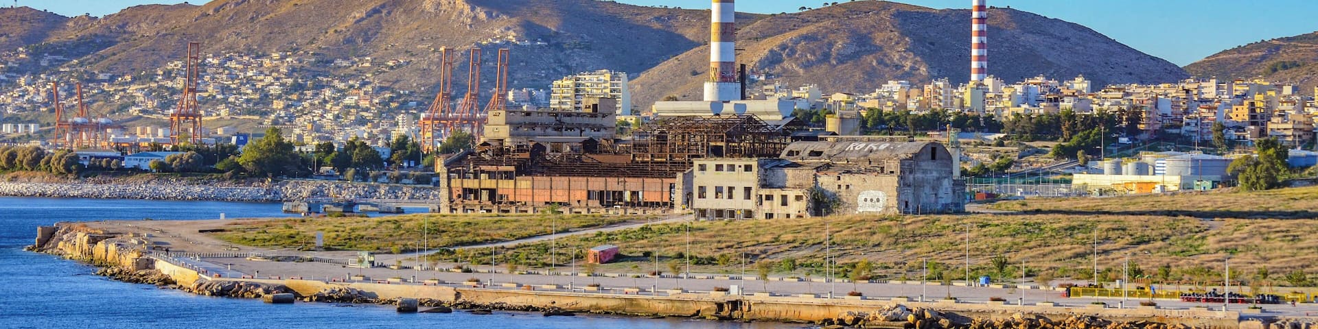 A coastal industrial area with a prominent tall chimney, a harbor, and a mountainous backdrop.