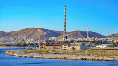 Zona costera industrial con una chimenea alta destacada, un puerto deportivo y un paisaje montañoso de fondo.