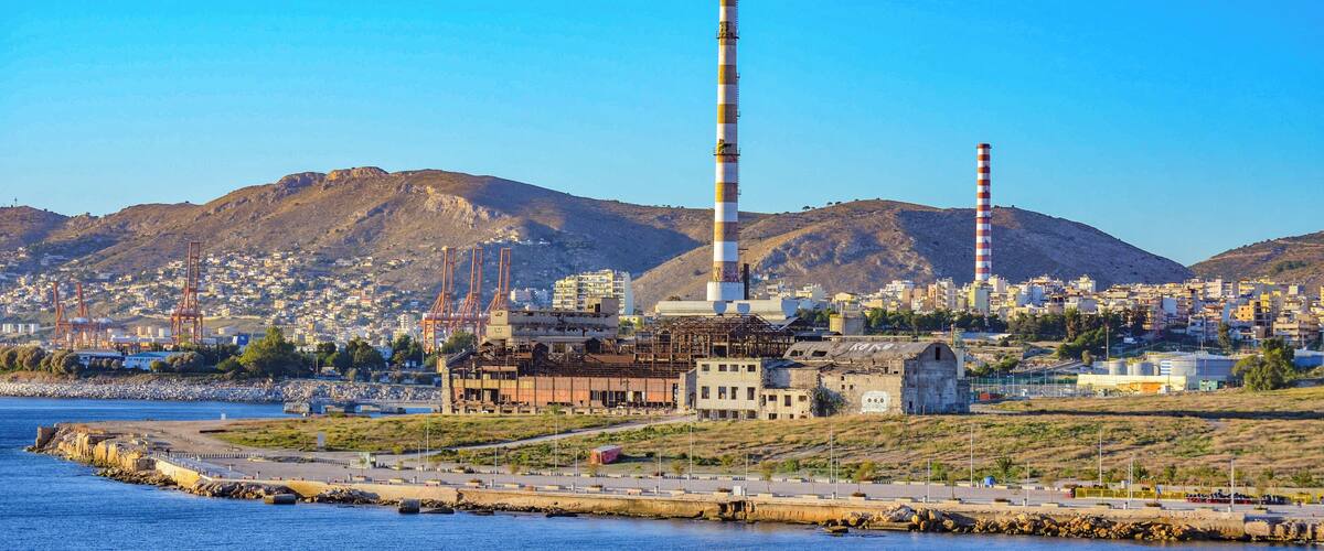 A coastal industrial area with a prominent tall chimney, a harbor, and a mountainous backdrop.