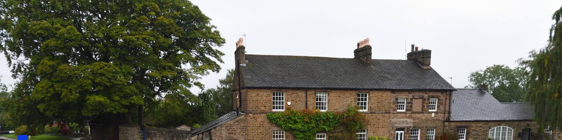 A narrowboat on a calm canal, a stone house with a garden, and a large tree in the background.