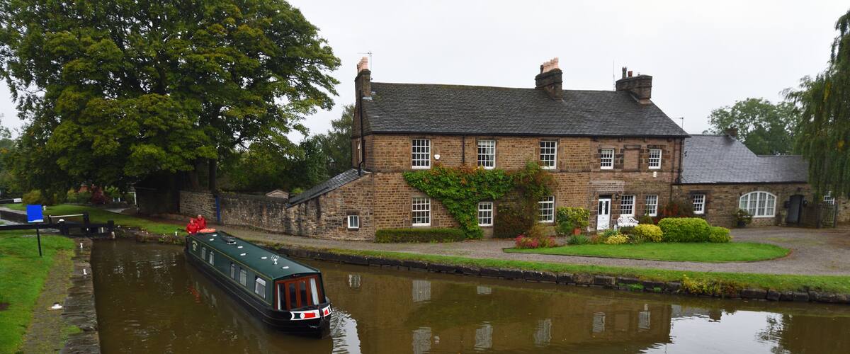 A narrowboat on a calm canal, a stone house with a garden, and a large tree in the background.