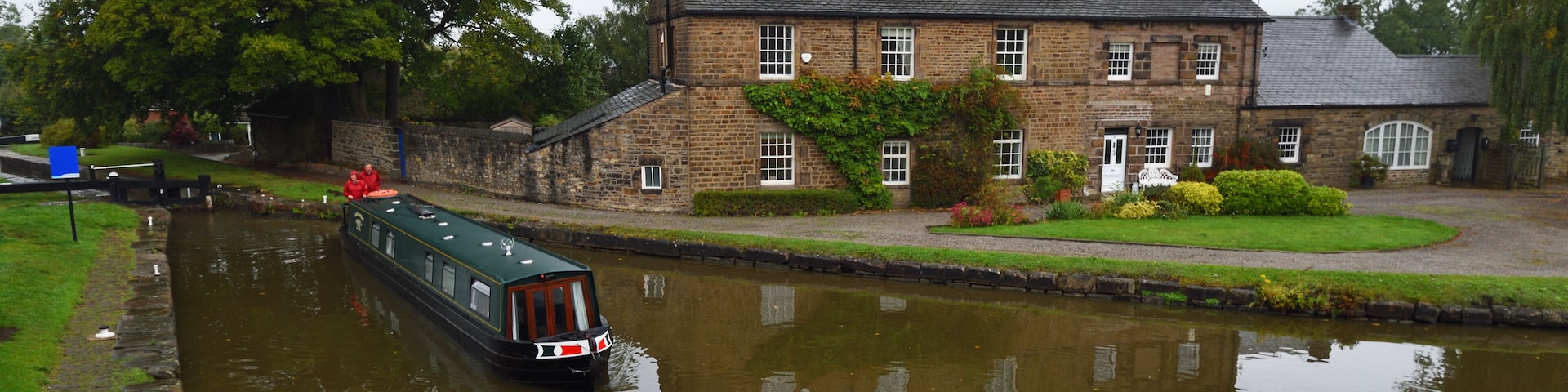 A narrowboat on a calm canal, a stone house with a garden, and a large tree in the background.