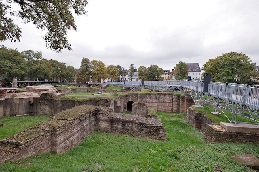 Ancient brick ruins with a grassy field, a bridge, and houses in the background.