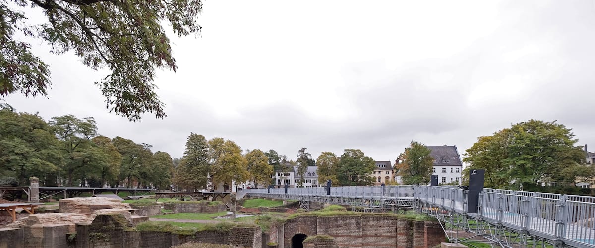 Ancient brick ruins with a grassy field, a bridge, and houses in the background.