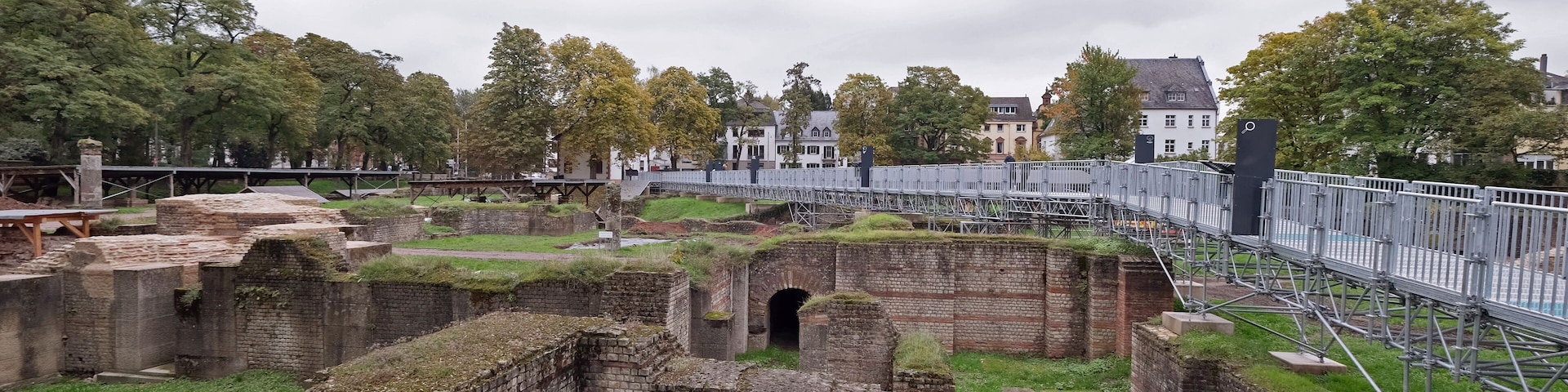 Ancient brick ruins with a grassy field, a bridge, and houses in the background.