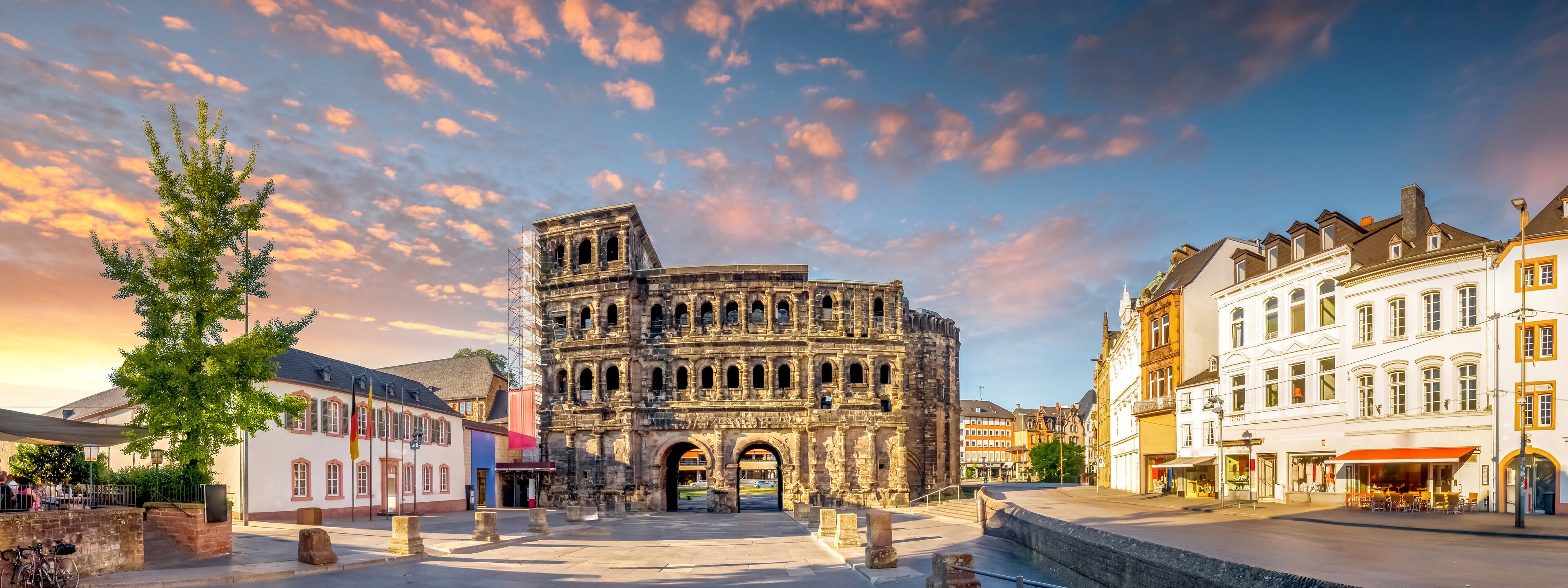 A historic stone structure with arches, surrounded by traditional European buildings and a clear sky with clouds.