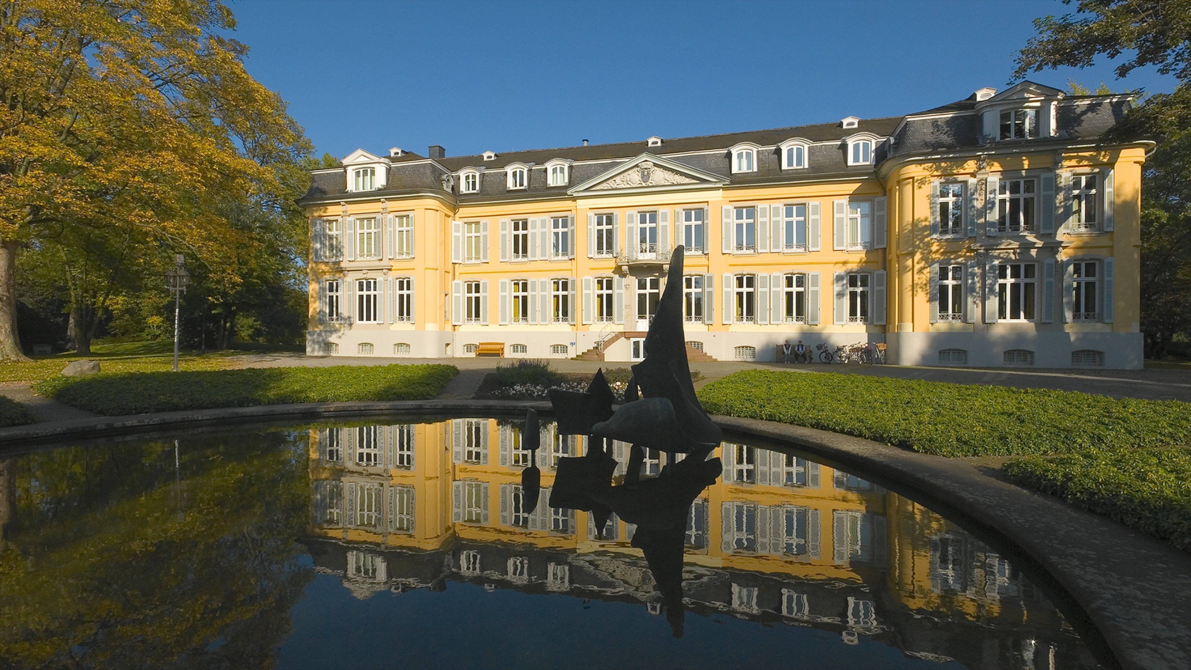 A large yellow building with white windows and a pond reflecting its image.