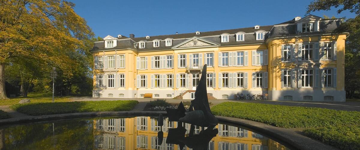 A large yellow building with white windows and a pond reflecting its image.