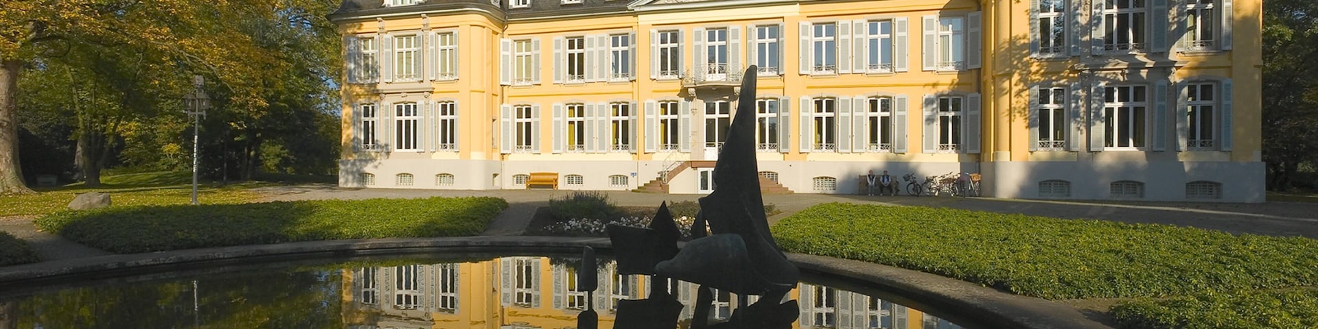 A large yellow building with white windows and a pond reflecting its image.