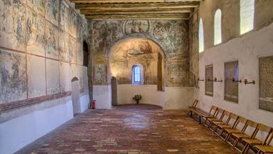 Interior com soalho de tijolos, bancos de madeira, frescos e uma janela central em arco.