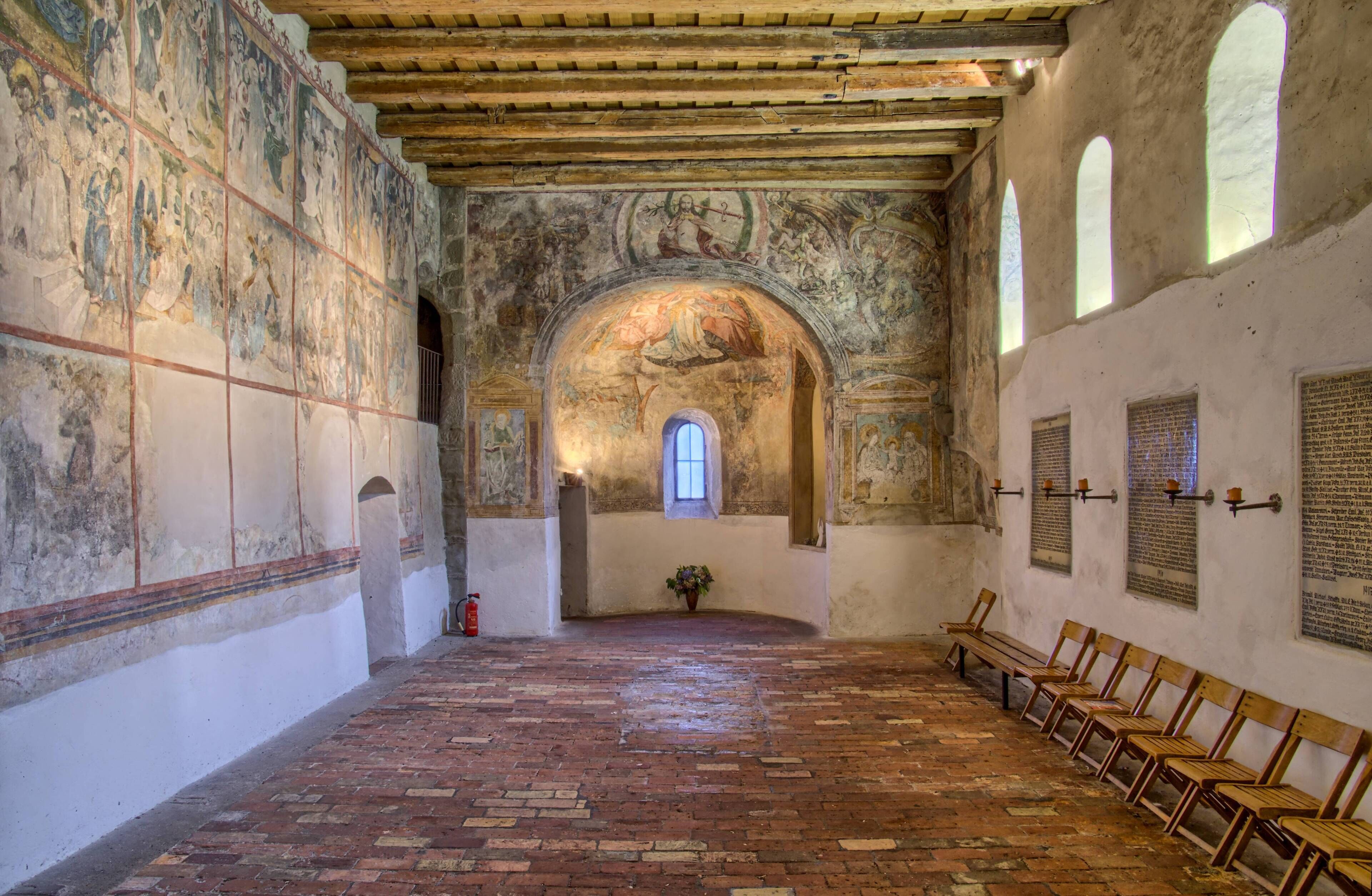 An interior with a brick floor, wooden benches, frescoes, and a central arched window.
