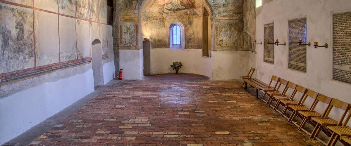 An interior with a brick floor, wooden benches, frescoes, and a central arched window.