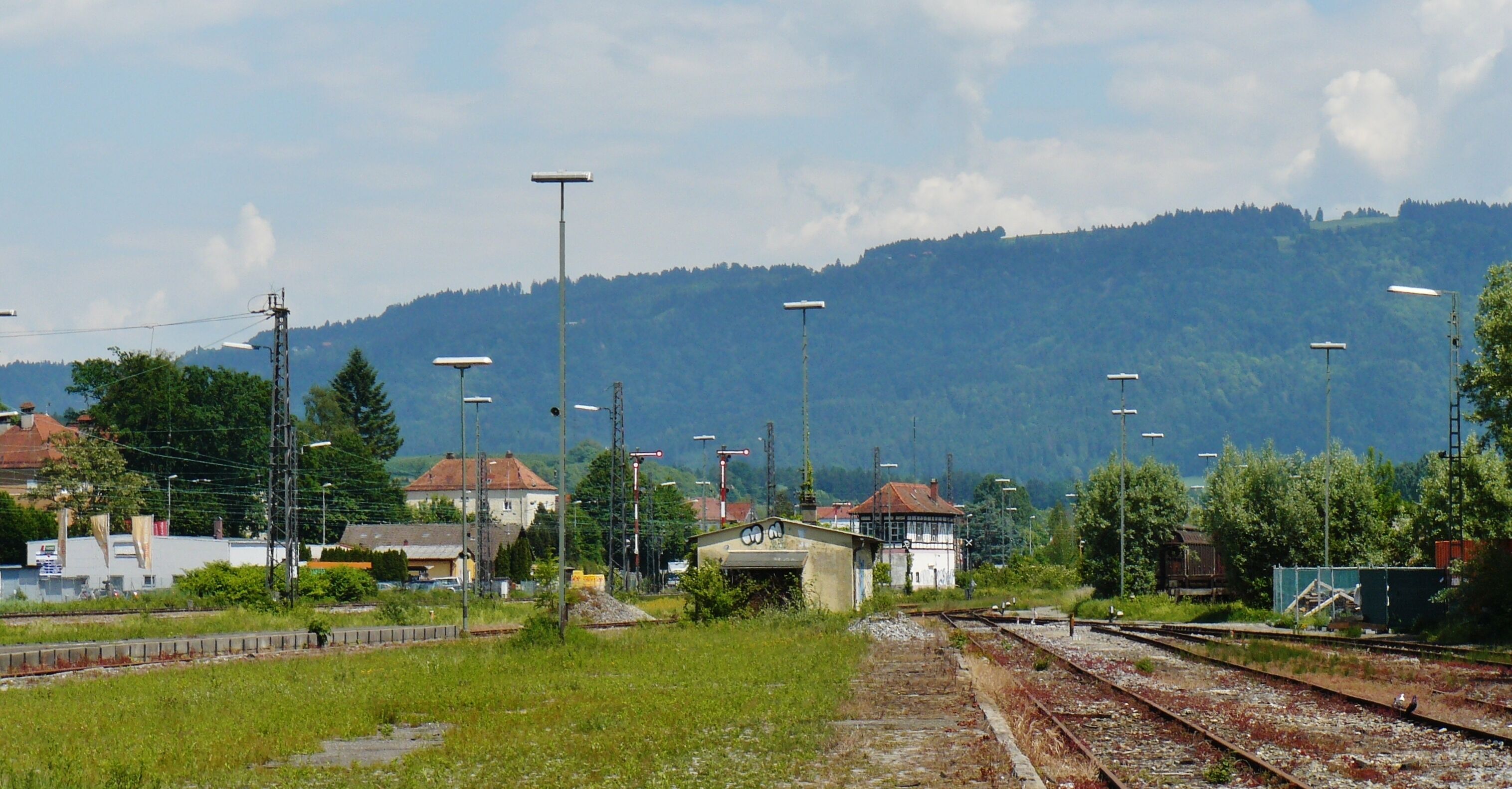 A railway crossing with multiple tracks, a small building, and a forested hill in the background.