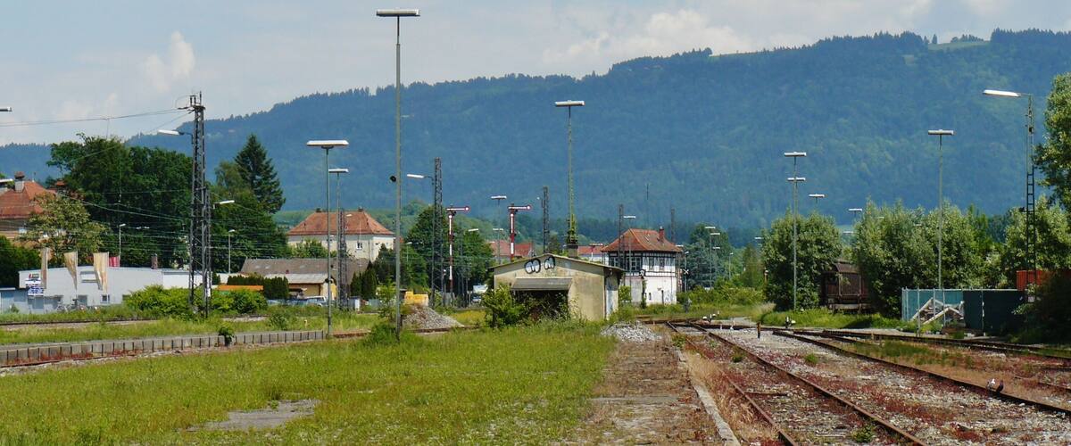 A railway crossing with multiple tracks, a small building, and a forested hill in the background.