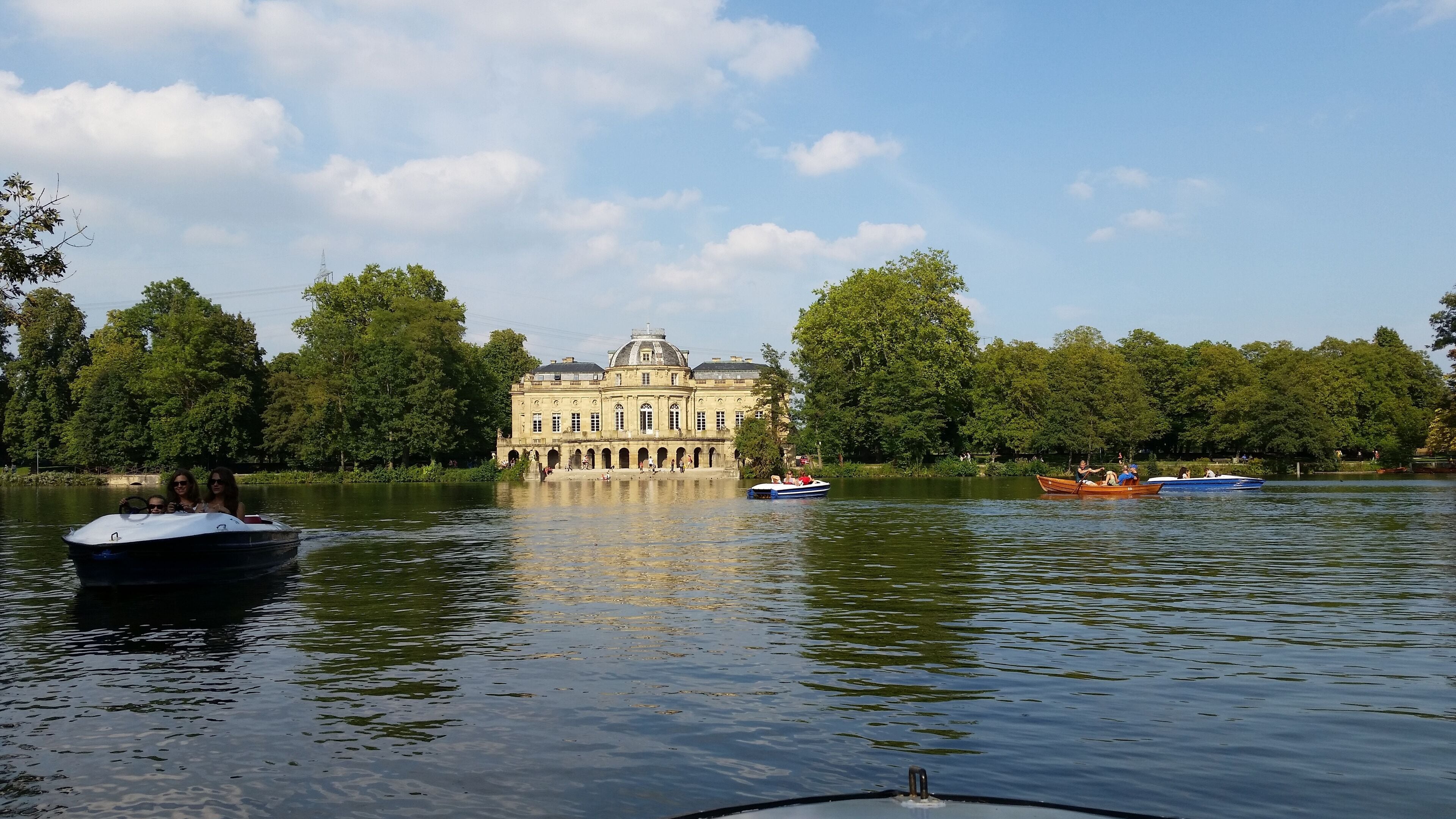 A historic building with a dome and arched windows, surrounded by a calm lake and lush trees.