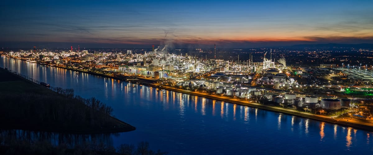 A cityscape at dusk with a river reflecting lights, industrial buildings, and a bridge.