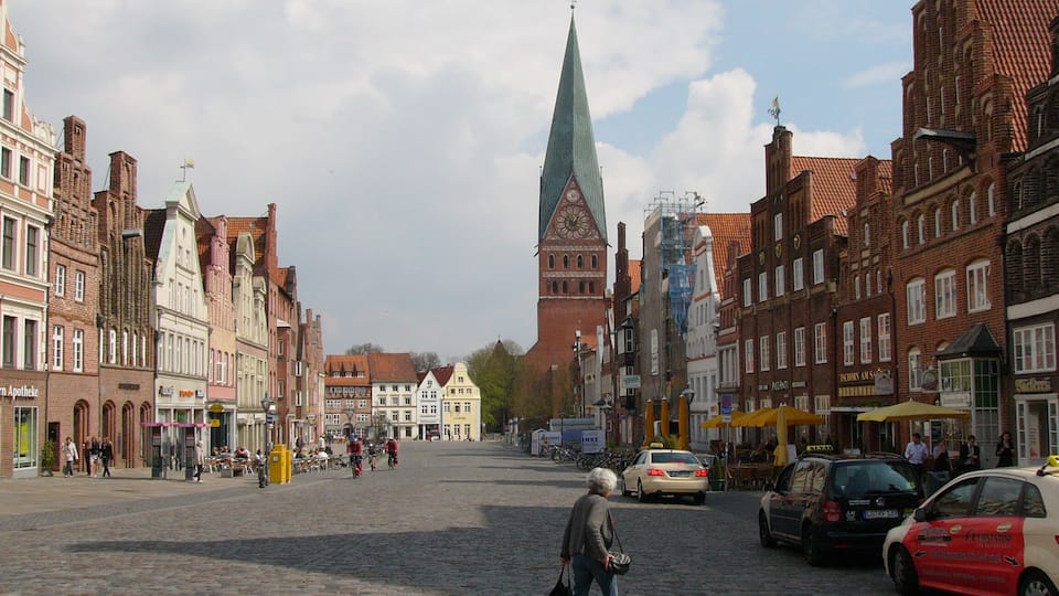 Eine gepflasterte Straße gesäumt von historischen Gebäuden, mit einem markanten Kirchturm und einigen parkenden Autos.