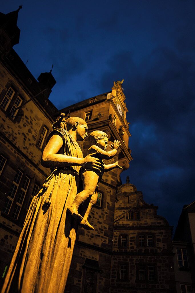 Eine Statue einer Frau, die ein Kind hält, vor einem historischen Gebäude mit einem Glockenturm.