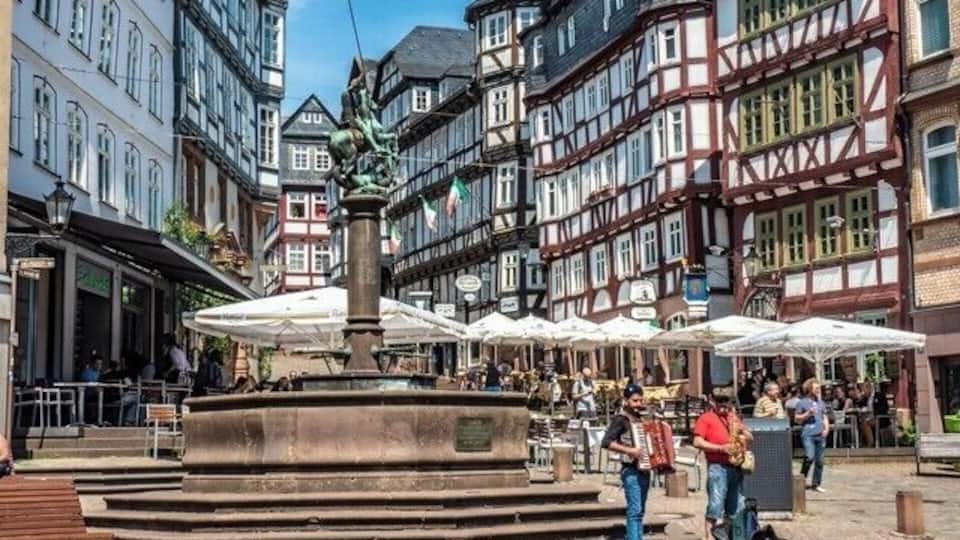 Ein historischer europäischer Stadtplatz mit einem zentralen Brunnen, traditionellen Fachwerkhäusern und Außenbereichen mit Sitzgelegenheiten.