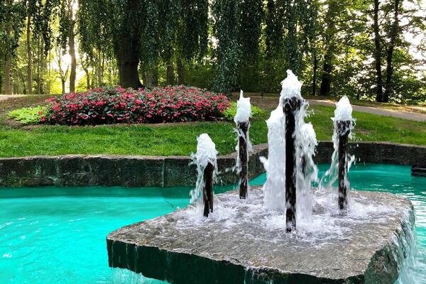 Ein Springbrunnen mit mehreren Wasserstrahlen in einem türkisfarbenen Becken, umgeben von üppigem Grün und einem Blumenbeet.