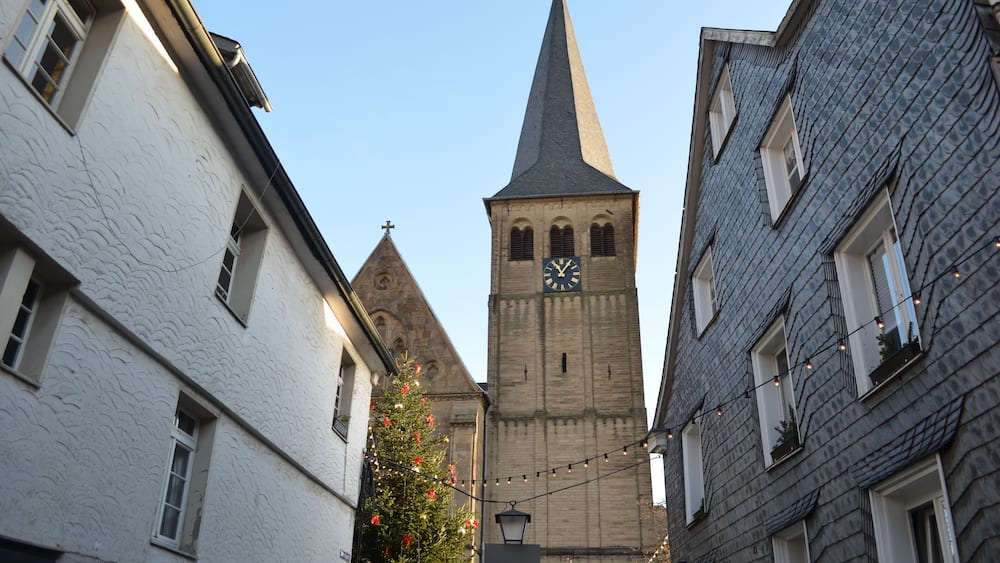 Eine schmale Straße mit einem Kirchturm, der eine Uhr zeigt, umgeben von Häusern mit strukturierten Fassaden.