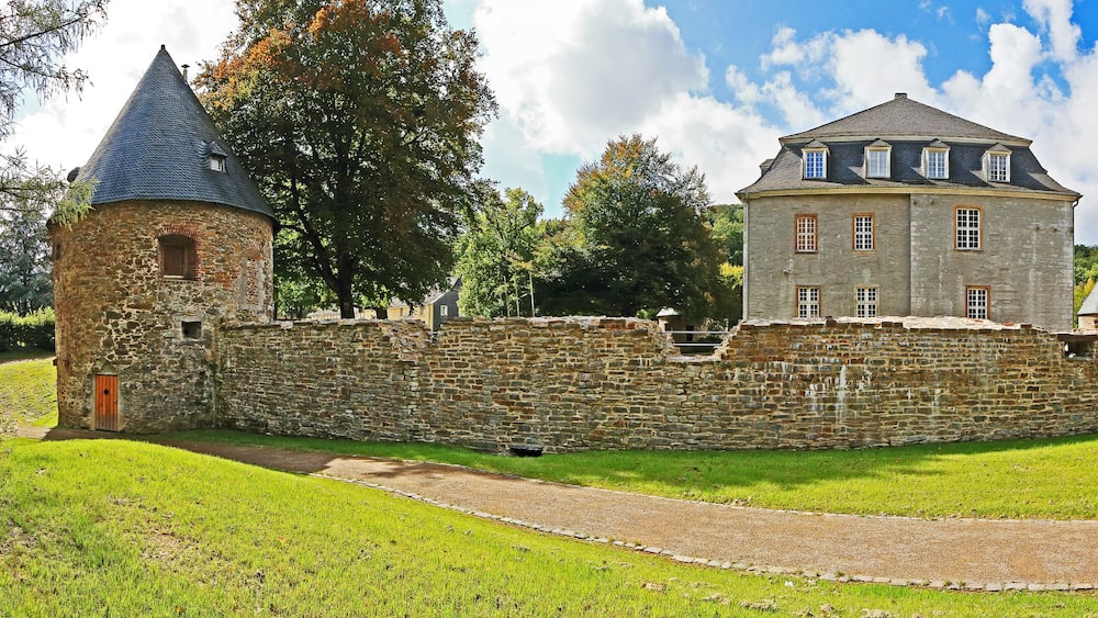 Un château historique en pierre, doté de deux tours et d’un bâtiment central, entouré d’un mur de pierre et d’une végétation luxuriante.