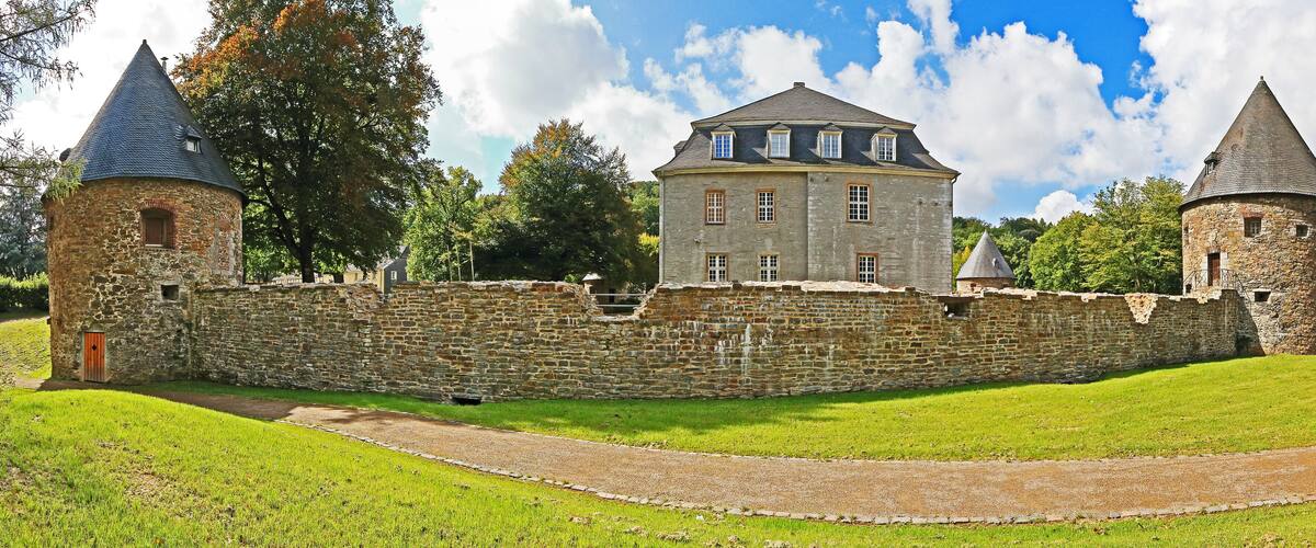 A historic stone castle with two towers and a central building, surrounded by a stone wall and lush green grass.
