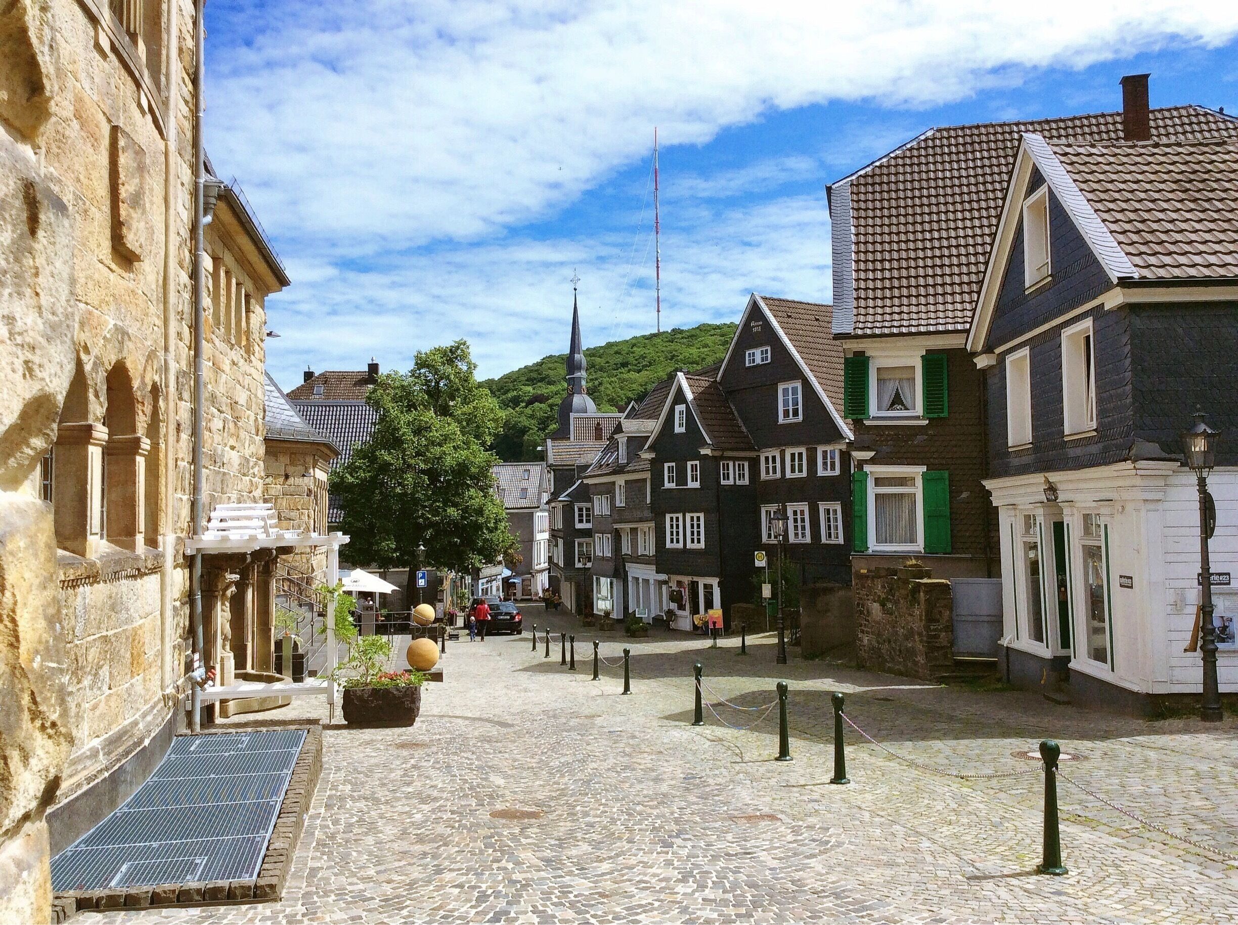 A cobblestone street lined with historic buildings, a church steeple, and a hill in the background.
