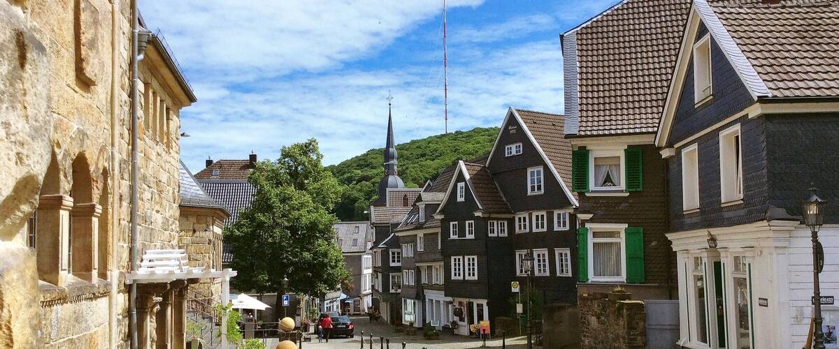 A cobblestone street lined with historic buildings, a church steeple, and a hill in the background.