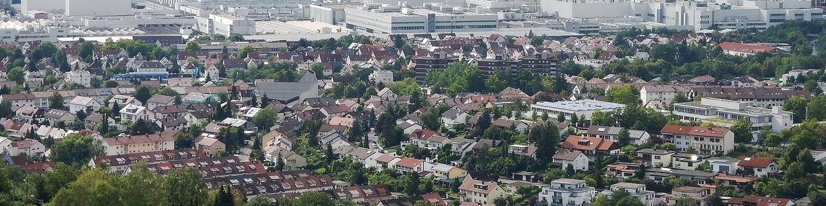 Un paysage urbain composé d’un mélange de bâtiments résidentiels et industriels, entouré de champs verdoyants et de collines lointaines.