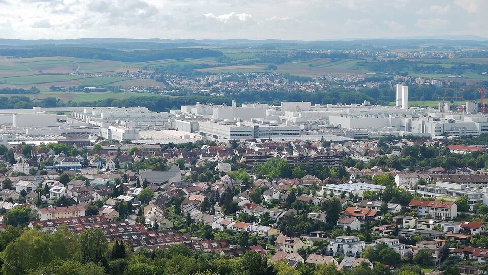 A cityscape with a mix of residential and industrial buildings, surrounded by green fields and distant hills.