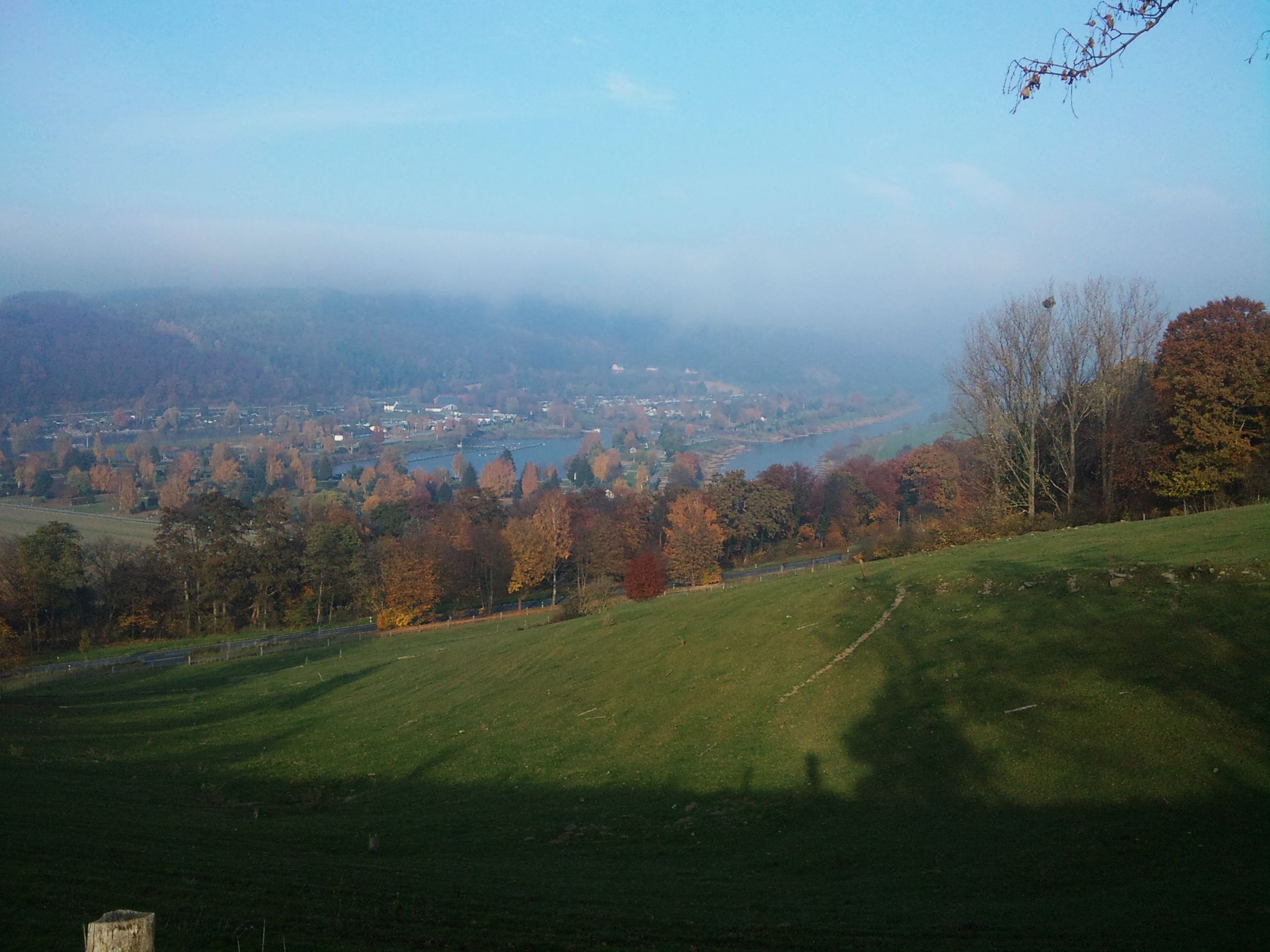 A panoramic view of a valley with a river, autumn-colored trees, and a distant town.
