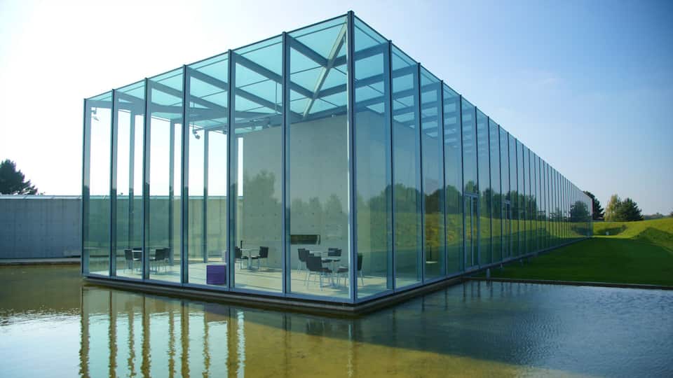 Una estructura moderna de vidrio con armazón metálico, ubicada sobre una superficie acuática reflectante.