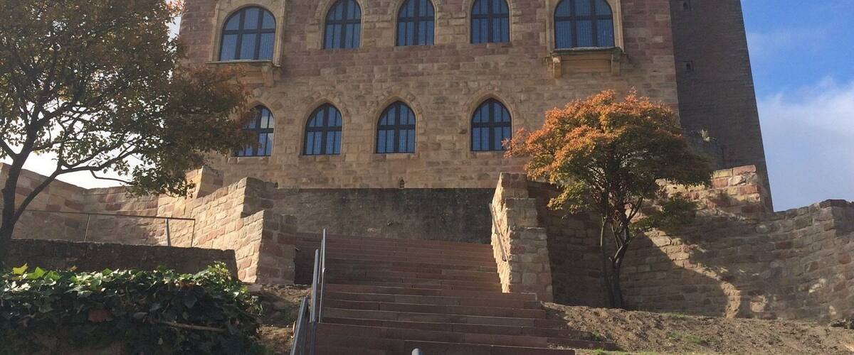 A historic stone building with arched windows and a tower, surrounded by a stone wall and steps leading up to it.
