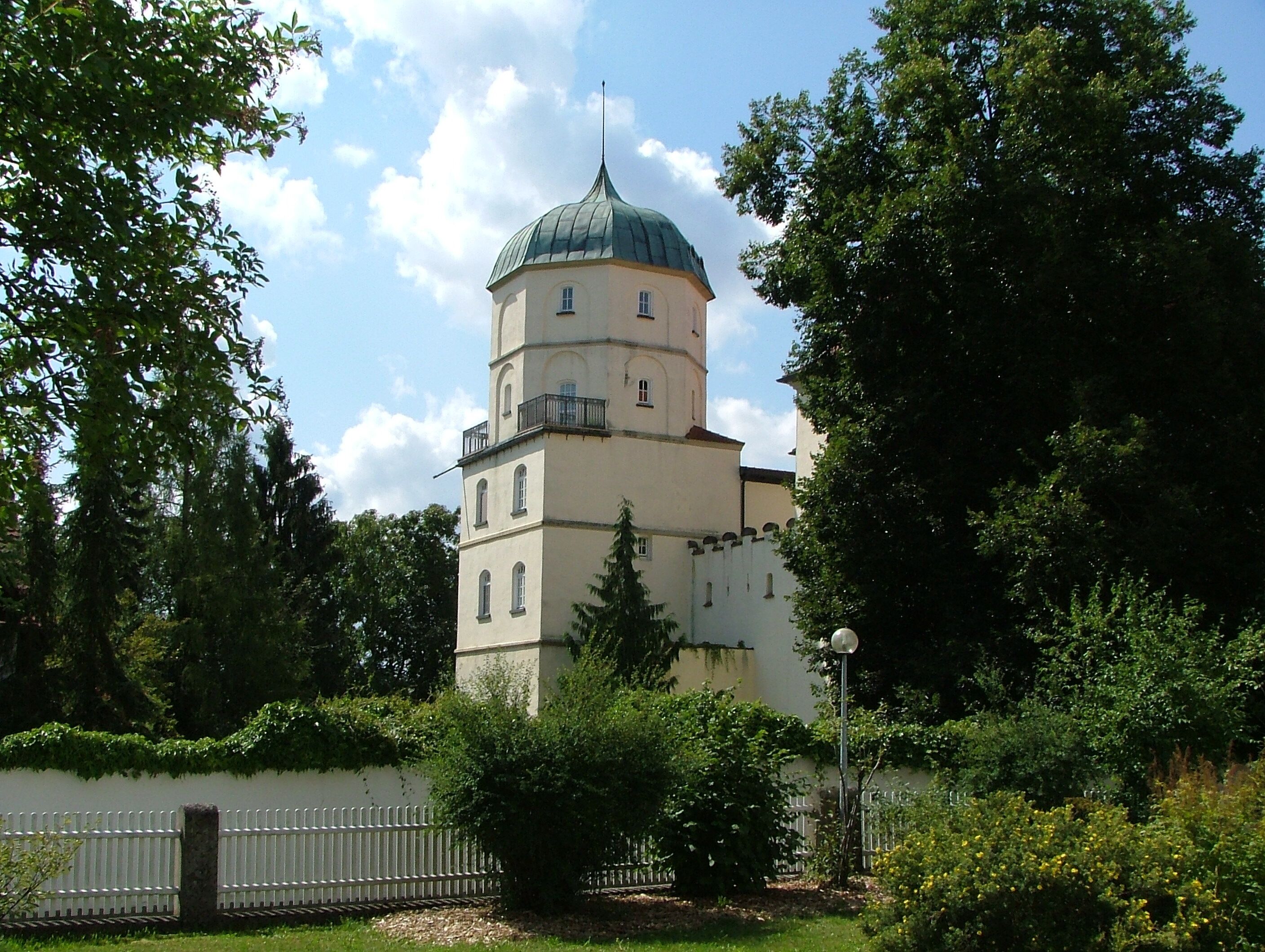 A historic building with a green dome and white walls, surrounded by lush greenery and a white fence.