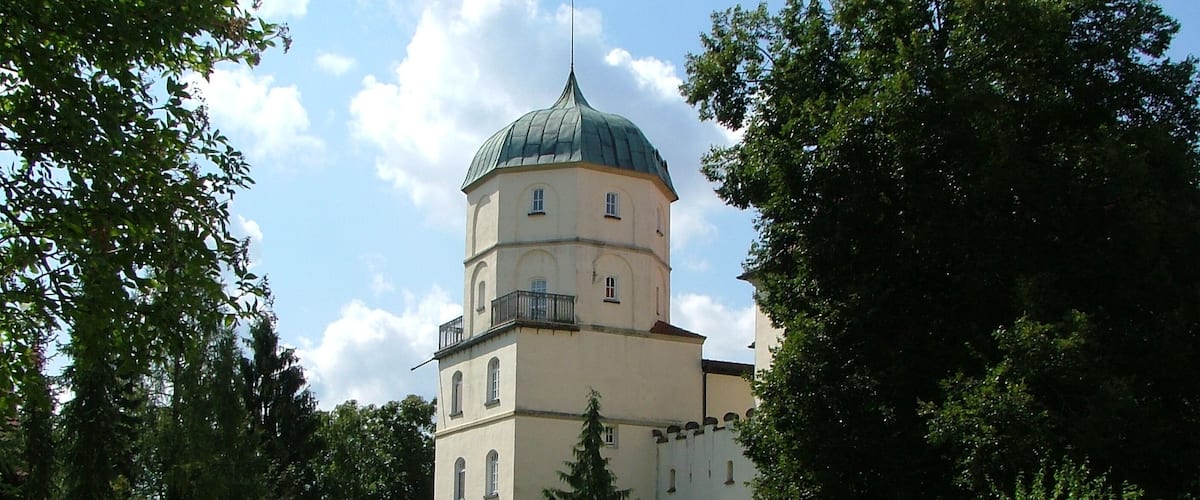 A historic building with a green dome and white walls, surrounded by lush greenery and a white fence.