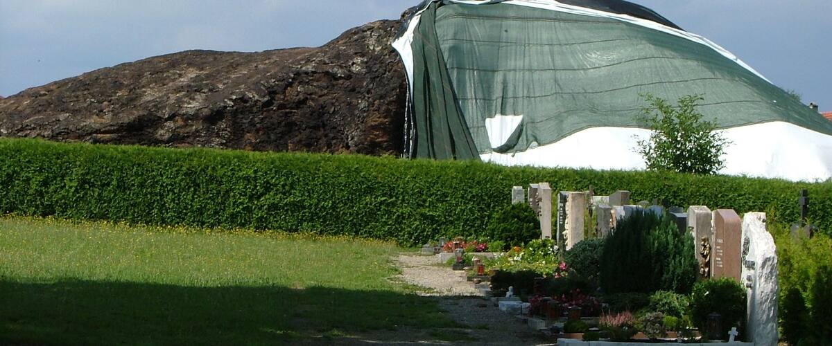 A large green tent-like structure with a dome on top, set against a rocky hill and a hedge.