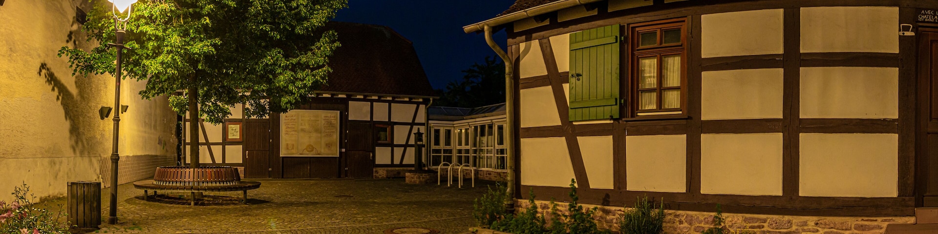 A cobblestone courtyard with traditional half-timbered houses, a bench, and a street lamp.