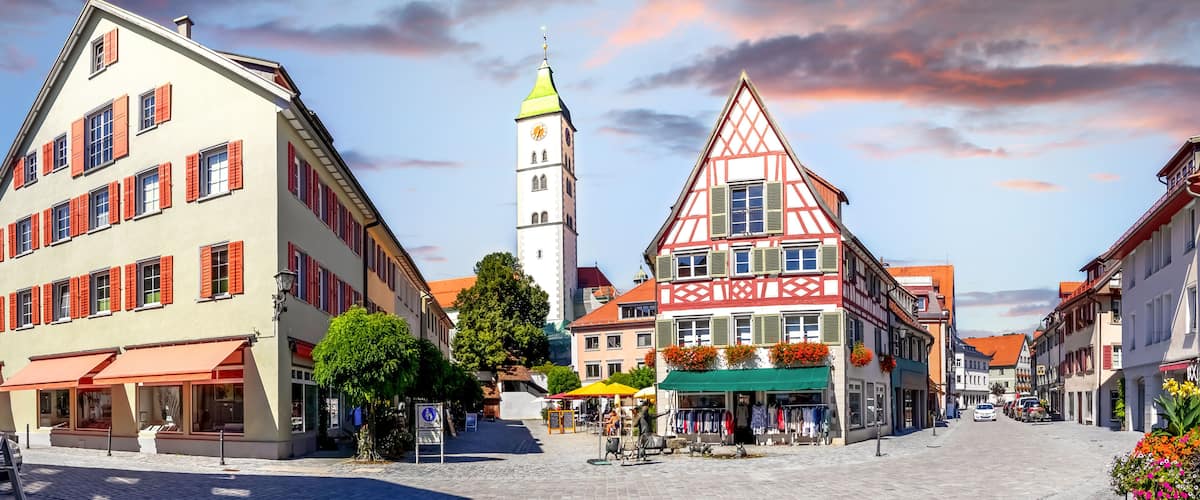 A European town square with traditional half-timbered buildings, a clock tower, and a cobblestone street.