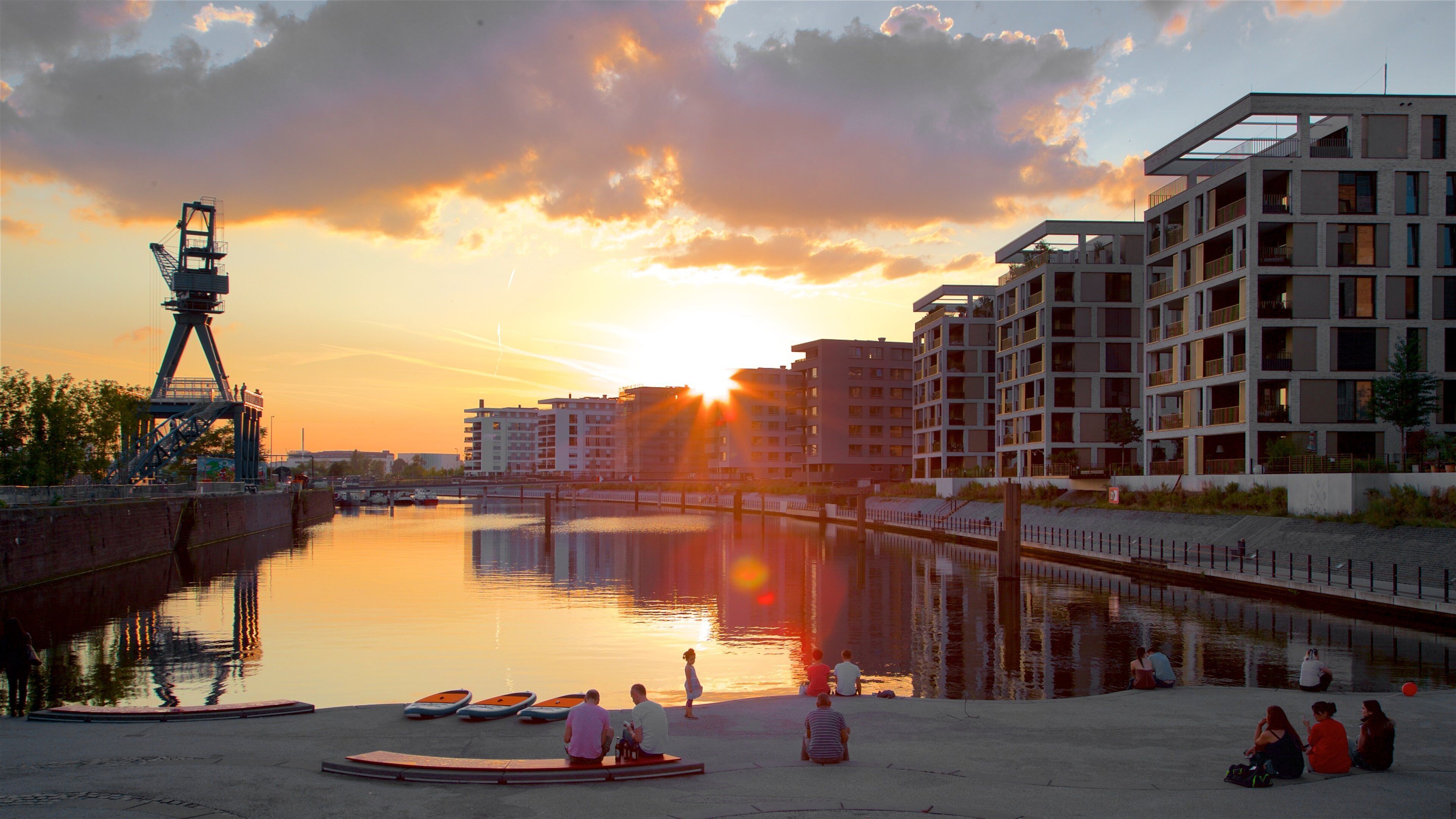 A sunset over a calm river with boats, a bridge, and modern buildings.