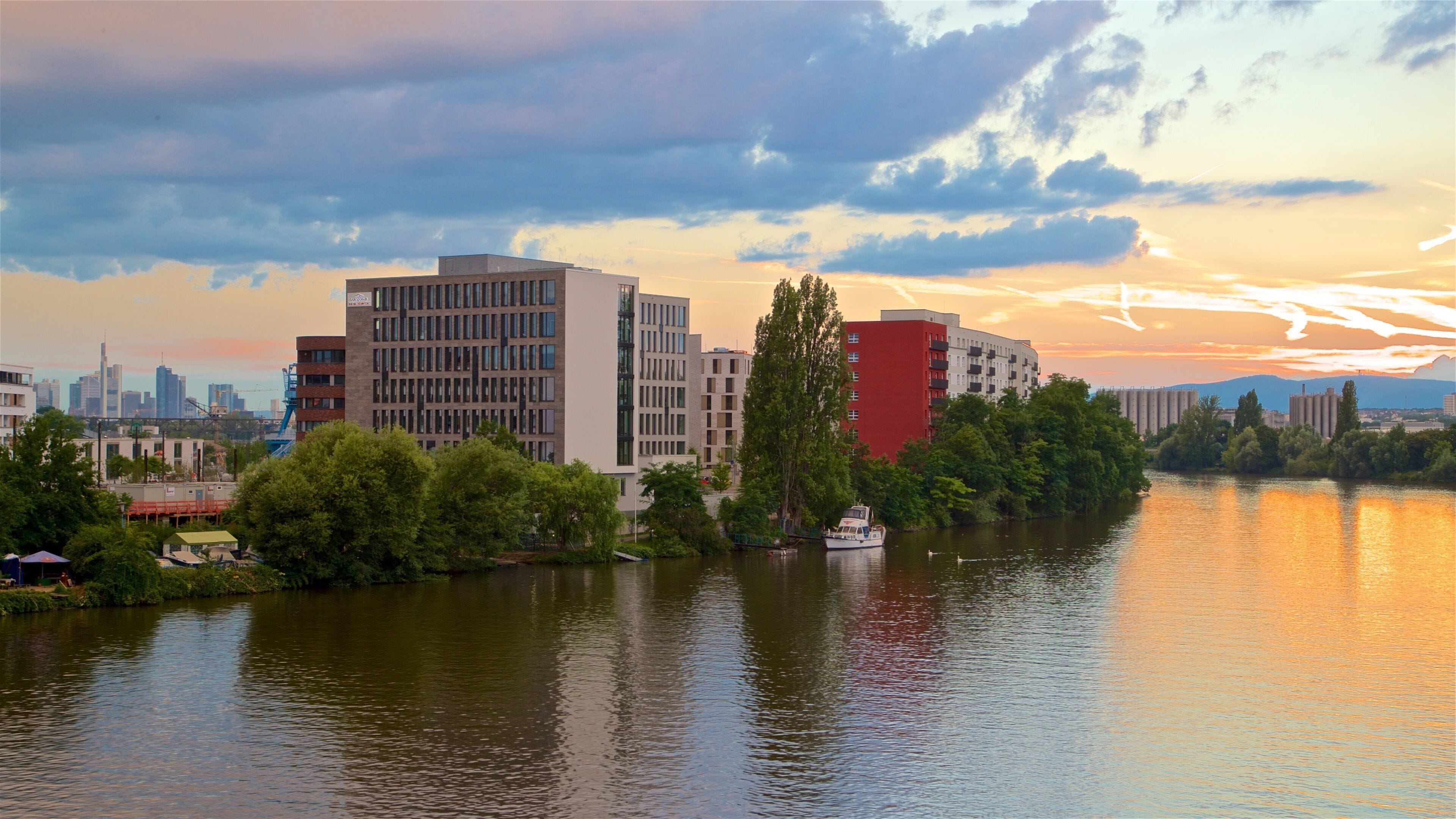 Un río con una barca, rodeado de edificios modernos y árboles, durante el atardecer.
