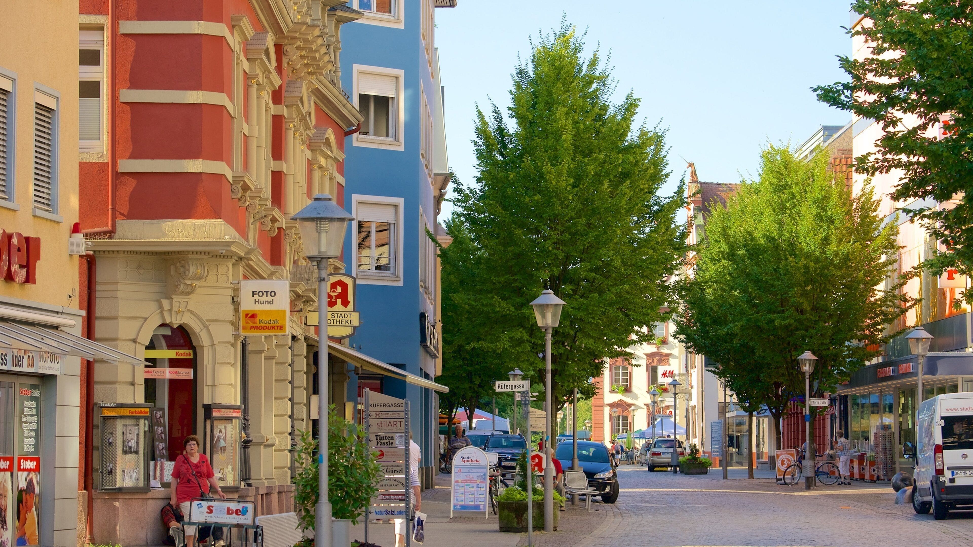A European street lined with colorful buildings, a street lamp, and a sign for a pharmacy.