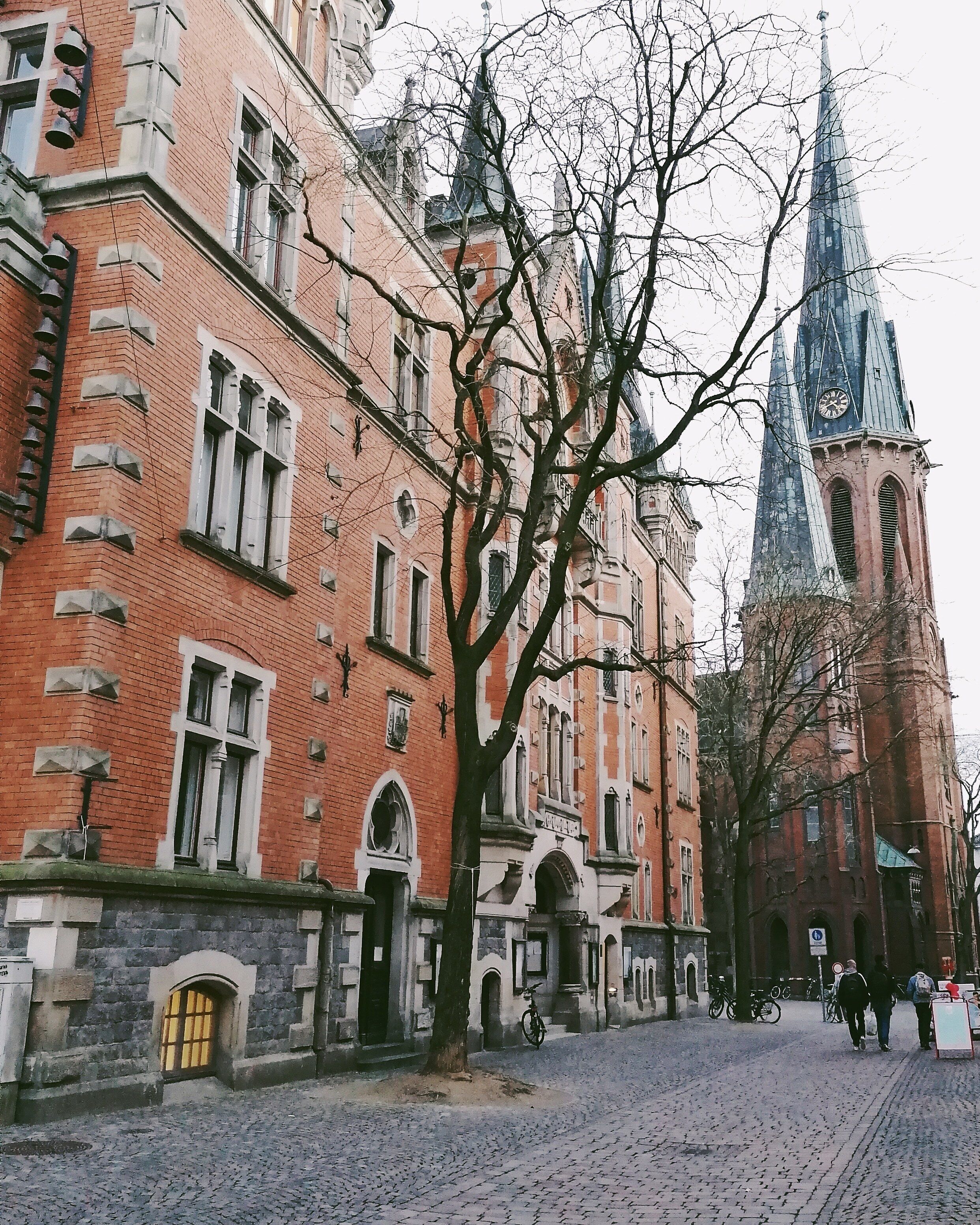 A cobblestone street lined with historic buildings, a prominent church tower, and leafless trees.