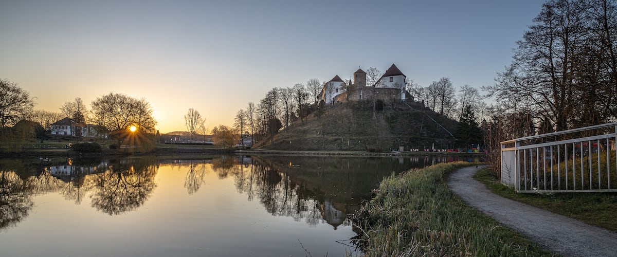 A calm lake reflecting a castle on a hill, surrounded by trees and a pathway.