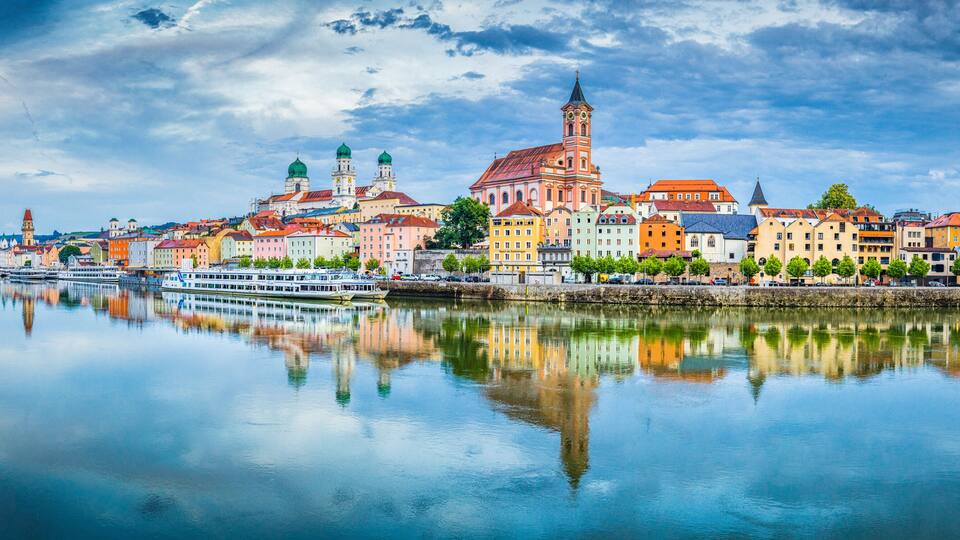 A river reflecting colorful buildings, a prominent church with a tall tower, and a bridge connecting the riverbanks.
