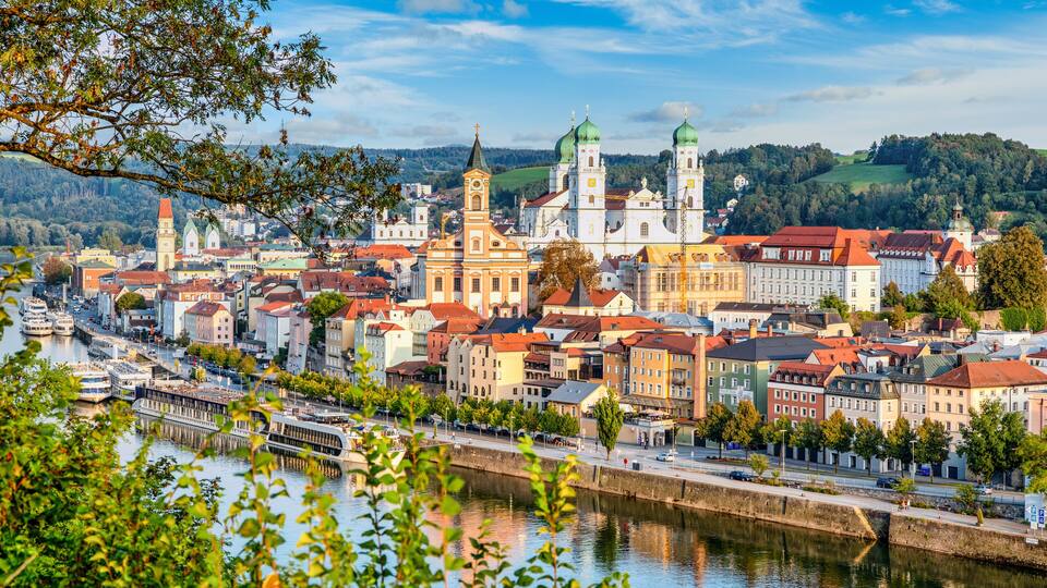 A riverside town with historic buildings, a prominent church with green domes, and a bridge.