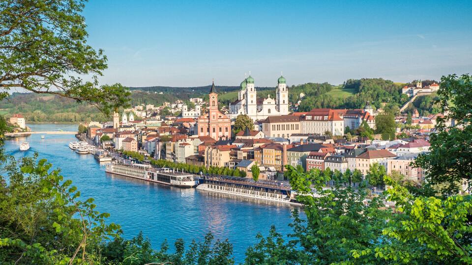 A riverside town with historic buildings, a prominent church with twin towers, and a river with boats.