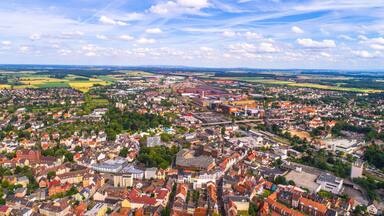 A panoramic view of a European town with a mix of historic and modern architecture, surrounded by green fields and a railway line.